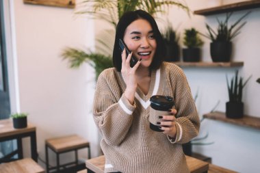 Kahveli neşeli Asyalı kız kafede akıllı telefondan konuşuyor. Genç, güzel, bin yıllık esmer kadın masada duruyor. Dinlenmek, boş zaman ve boş zaman kavramı. Modern kadın yaşam tarzı