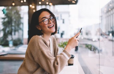 Kafede kameraya bakan akıllı telefonlu Asyalı bir kız. Bin yıllık neşeli esmer kadın pencerenin yanındaki masada gözlük takıyor. Dinlenmek, boş zaman ve boş zaman kavramı. Modern kadın yaşam tarzı