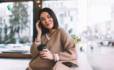Kahve içen Asyalı bir kız ve kafede kameraya bakıyor. Genç gülümseyen milenyum esmer kadın pencerenin yanındaki masada duruyor. Dinlenmek, boş zaman ve boş zaman kavramı. Modern kadın yaşam tarzı