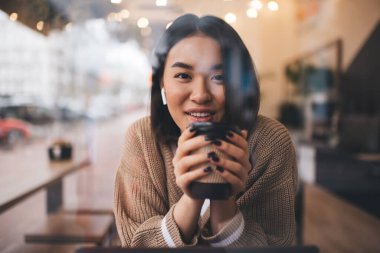 Cafe 'de çalışırken gülümseyen Asyalı kızın portresi. Genç milenyum esmer kadın kulaklık takıyor ve kameraya bakıyor. Serbest ve uzaktan kumandalı. Modern kadın yaşam tarzı