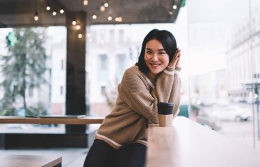 Gülümseyen Asyalı kız dinleniyor ve kafede kameraya bakıyor. Genç milenyum esmer kadın elinde kahveyle pencerenin yanında duruyor. Dinlenmek, boş zaman ve boş zaman kavramı. Modern kadın yaşam tarzı