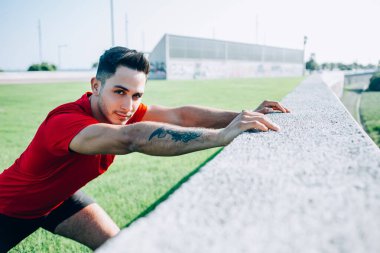 Spor kıyafetlerinde kendine güveni tam bir etnik adam koşuya hazırlanıyor. Stadyumdaki duvara yaslanıp kameraya bakıyor.
