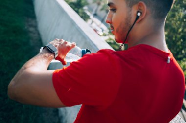 Su şişesi ile beyaz erkek antrenör spor amaçlarını analiz etmek için bilek saatine bakıyor. Elektronik kulaklıklı genç sporcu, sabah egzersizi için müzik listesi seçiyor.