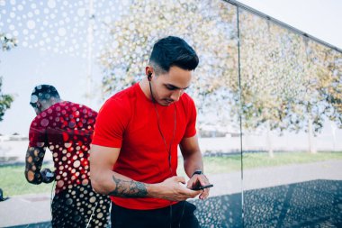 Kırmızı tişörtlü genç kaslı, güneşli bir günde sokakta duran kulaklıklı cep telefonuyla müzik dinliyor. 