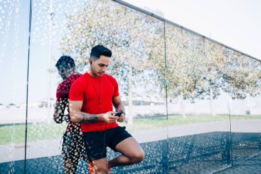 Kırmızı tişörtlü genç atletik kaslı. Güneşli bir günde sokakta duran kulaklıklarla cep telefonundan müzik dinliyor. 