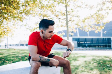 Kırmızı tişörtlü genç, kaslı atletik adam güneşli bir günde şehirde spor sahasında bilek saatlerini kontrol ediyor.