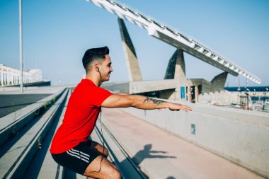 Kulaklıklı, sportif yaz kıyafetleri giymiş, gündüzleri merdivenlerde çömelme egzersizi yapan neşeli genç bir erkek.