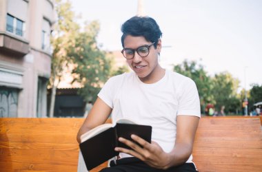 Görsel düzeltme için moda gözlüklü mutlu Latin öğrenci olumlu edebiyat romanı okumak için boş vakitlerin tadını çıkarıyor, tasasız erkek blogcu ilginç bir yayın yaparken plan yapıyor.