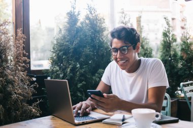 Neşeli erkek bilgi işlem uzmanı hareketlilik bildirimleri okuyor ve internet bağlantısı iyi olan bir kafede oturmuş gülümsüyor gözlüklü adam akıllı telefon aracılığıyla internet mesajlaşmasından hoşlanıyor.