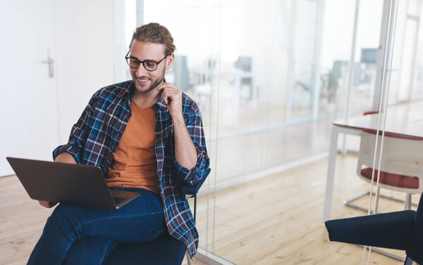 Millennial caucasian businessman using and watching on latop during work. Concept of modern successful man. Young smiling male entrepreneur in glasses sitting on office chair in open space office