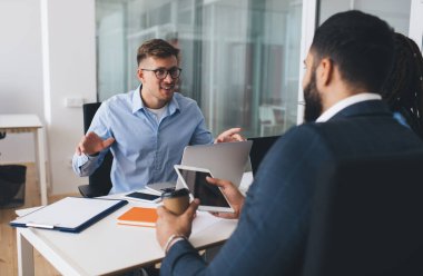 Optik gözlük takan Kafkas erkek çalışanı, masa başında çalışma saatlerinde modern teknoloji ve iş adamlarıyla yapılan brifingleri tartışan profesyonel meslektaşlarıyla iletişim kuruyor.