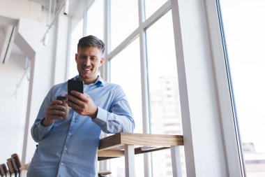 Kafkasyalı bir adam cep telefonu uygulaması üzerinden sosyal medyaya bağlanırken içerik metinlerine göz atmak için kahve içiyor, cep telefonu teknolojisi sohbet ve mesajlaşmalarıyla gülümseyen erkek blogcu