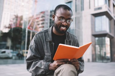Gözlüklü neşeli erkek öğrenci dışarıda öğrenim görürken edebiyat kitaplarından bilgi topluyor. Bilgi için ders kitabı olan mutlu Afro-Amerikan hipster adam notları analiz ediyor.
