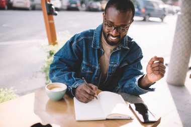 Not defterine ödevini yazan neşeli hippi adam kaldırımdaki kafede eğitim zamanının keyfini çıkarıyor, klasik gösterilerde mutlu Afrikalı Amerikalı erkek öğrenci tasarım projesi için skeç hazırlıyor.