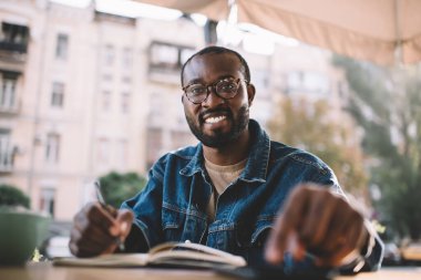 Eğitim kitabı olan Afro-Amerikan erkek öğrenci kaldırım kafesinde öğrenim ve öğrenim görüyor, klasik gözlüklü neşeli hippi çocuk sınavlara hazırlanıyor ve açık havada gülümsüyor.