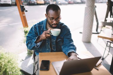 Kahve fincanı ile serbest çalışan yetenekli bir erkek netbook cihazıyla sokak kafesinde webinar izlerken klasik göz giysisi okuma içeriği yayınlayan bin yıllık koyu tenli bir adam.