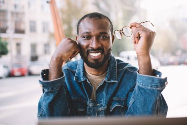 Optik gözlüklü neşeli hipster adamın portresi kaldırımdaki kafede boş zamanlarında kameraya gülümseyen, elinde şık gözlük olan neşeli Afro-Amerikan adam.