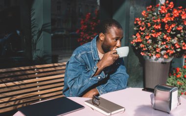 Kafede dinlenirken ve beklerken kafein içeceği içerken koyu renkli kot ceketli koyu tenli erkek, kapalı dizüstü bilgisayarı ve ders kitabı olan Afro-Amerikan dijital göçebe açık havada kahve tadı alıyor.