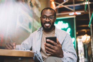 İçerde akıllı telefon uygulaması kullanırken kameraya gülümseyen optik gözlüklü neşeli erkek öğrenci portresi, modern mobil cihazlı mutlu Afrikalı Amerikalı hipster adam cep telefonunun keyfini çıkarıyor.