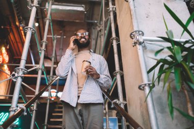 Optik gözlüklü neşeli siyahi hippi arkadaş cep telefonuyla gülümsüyor. Boş zamanlarında sohbet ediyor. Afro-Amerikan bir adam, kahve saatinde telefon uygulaması ile telefon görüşmesi yapıyor.