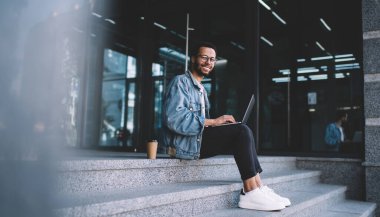 Optik gözlüklü neşeli hipster adamın portresi kameraya göz düzeltmesi sağlıyor netbook 'ta mesafesini tamamladığı için gülümsüyor şehir merdivenlerinde dizüstü bilgisayarla poz veren mutlu erkek blogcu.