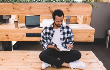 Eğitim kitabı ve boş dizüstü bilgisayarı olan Millennial erkek öğrenci. Arka planda kopyalama alanı olan bir bilgisayar. Öğrenim görürken notlar alıyor.