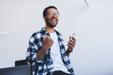 Optik gözlüklü neşeli hippi iş fırsatları hakkında mobil haberler aldı. Ofis içi akıllı telefon taraması sırasında mutlu bir erkek çalışanın online hedefini kutlaması.