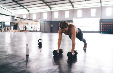 Sportif giyinen güçlü bir kadın spor salonunda egzersiz yaparken şınav çekiyor. Eşofmanlı kararlı bir kadın antrenör. Dambıllar vücudunu fiziksel çabayla formda tutuyor.