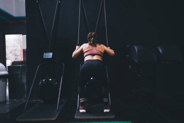 Profesyonel genç bayan atlet spor salonunun içinde çekiş makineleri üzerinde sıkı çalışıyor, formda bir kızın sağlıklı yaşam tarzını arka planda tutarken kapalı alanda ağırlık kaldırmak için simülatör kullanıyor.