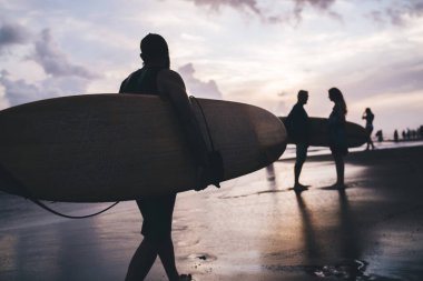 Okyanusta amatör gezintiler için elinde profesyonel sörf tahtası tutan tanınmayan bir adamın silueti, kıyı şeridinde yürüyen erkek sörfçülerin arka görüntüsü Phuket, Bali Endonezya 'da aşırı derecede eğlenen
