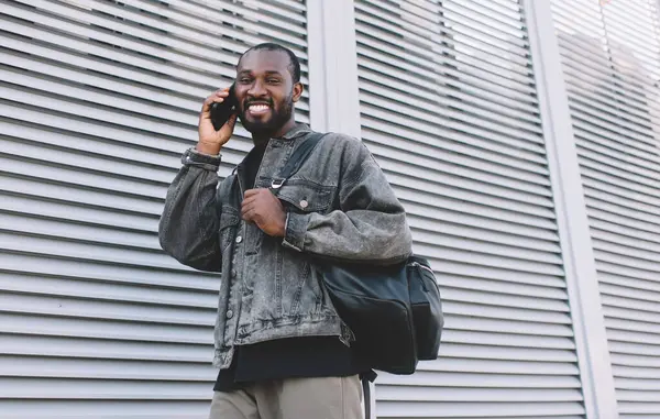 Seyahat sırt çantalı esmer bir adam uluslararası telefon görüşmesinden memnun. Şehirde geziniyor, neşeli hipster adamın portresi. Akıllı telefon uygulaması ile aramak için 4G kablosuz kullanıyor.