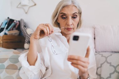 Odaklanmış beyaz kadın evdeki kanepede akıllı telefondan bir şey izliyor. Ev istirahatı ve boş zaman kavramı. Gri saçlı gözlüklü kadın. Modern geniş dairenin içi.
