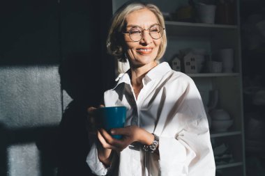 Bir fincan çay ya da kahveyle gülümseyen Kafkas iş kadını seramik ürünlerle dolu karanlık ev sanat atölyesine bakıyor. Küçük işletmeler ve girişimcilik. Modern başarılı kadın