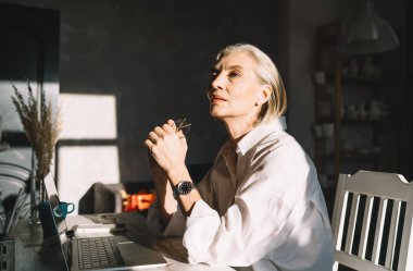 Üst düzey Kafkasyalı iş kadını, karanlık ev sanatları stüdyosunda güneşli ışıklandırmalı bir masada çalışırken bir şeyler düşünüyor. Küçük işletmeler ve girişimcilik. Modern başarılı kadın
