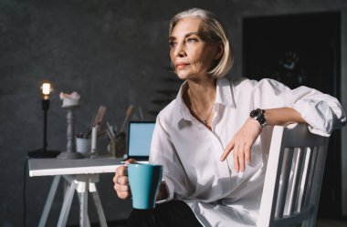 50 yaşında, beyaz bir iş kadınına odaklanmış, elinde bir fincan kahveyle sandalyeye oturmuş, başka tarafa bakıyor. Küçük işletmeler ve girişimcilik. Modern başarılı kadın. Güneşli ışıklandırmalı karanlık sanat stüdyosu