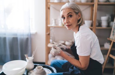 Cidden 50 yaşında, beyaz kadın heykeltıraş kil çömlek yapıp kameraya bakıyor. Küçük işletme ve girişimcilik kavramı. Ev hobisi, eğlence ve eğlence. Sanat stüdyosunda çalışan bir kadın.