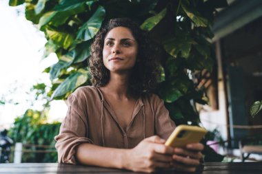 Kafe terasında dinlenen modern hücresel cihazlı düşünceli kadın blogcu yayınlamak için web içeriğini düşünüyor, kıvırcık saçlı düşünceli hippi kız dijital cep telefonu tutuyor.