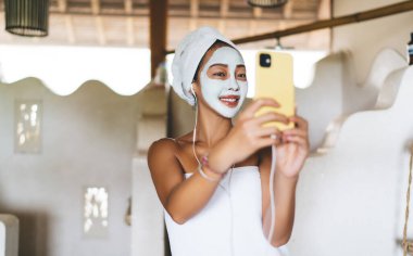 Mutlu Asyalı kadın, evde banyo yaparken maske takarak güzellik videosu çekiyor. Dijital cep telefonuyla ön kamerayı kullanıyor, kulaklıklı neşeli bir kadın selfie çekiyor.