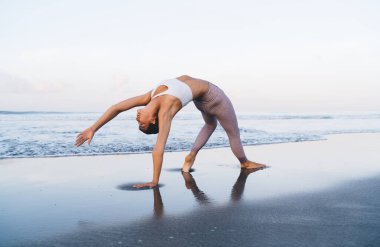 Eşofmanlı motive bir kadın yoga pozisyonunda duruyor. Deniz kıyılarında nefes alıyor ve eğleniyor. Aktif kadın yogası yapıyor.