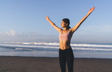 Siyah taytlı ve pembe tişörtlü çekici kadın yogi kıyı şeridinde güneş ışığını seyrediyor spor giysili beyaz kadın canlılık ve sağlığa karşı ısınıyor.