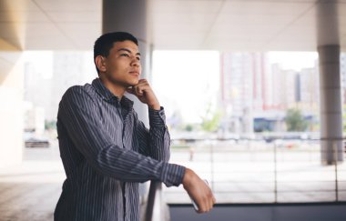 Asyalı genç bir delikanlı bir şey düşünüyor ve dışarı bakıyor. Uzaktan kumanda ve serbest çalışma kavramı. Modern, başarılı bir adamın fikri. Ofis binasının balkonunda bir erkek. Güneşli bir gün