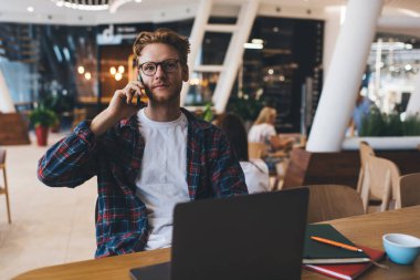 Genç, beyaz bir adam, bilgisayarını açmış, akıllı telefondan konuşuyor. Çevrimiçi eğitim ve iş kavramı. Modern, başarılı bir adamın fikri. Ofiste çalışan ya da ders çalışan gözlüklü ciddi bir adam.