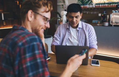 Asyalı erkek arkadaşı dizüstü bilgisayarda yazarken beyaz iş adamı akıllı telefondan izliyor. İş birliği ve takım çalışması. Modern başarılı adamlar. Geniş ofisteki masada çalışan gençler.