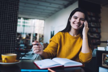 Genç, pozitif bir kadın cep telefonuyla konuşuyor ve gülümsüyor. Modern kitap kafeteryasında açık bir defter ve bir fincan kahveyle oturuyor.