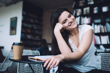Kahve molası sırasında masada oturan beyaz tişörtlü, gülümseyen bayan ve başka tarafa bakıp buluşmayı beklerken kahkahalar atan.