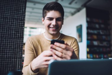 Gündüz vakti kafede dizüstü bilgisayarla oturup cep telefonundan iyi haberler okurken gülümseyerek mutlu genç bir erkek.