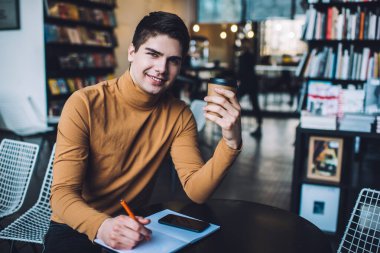 Pozitif genç erkek elinde bir bardak sıcak içecekle yaratıcı bir yerde oturmuş kameraya bakıyor.