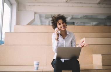 Bilgisayarlı, akıllı telefondan konuşan bin yıllık beyaz bir kız. Modern başarılı kadın kavramı. Genç gülümseyen esmer kadın, çalışma odasında, ahşap bankta, kahve fincanıyla oturuyor.