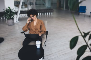 Milenyum Avrupalı iş kadını akıllı telefondan konuşur ve iş sırasında dizüstü bilgisayarda izler. Modern başarılı kadın kavramı. Ofisteki koltukta oturan, gözlüklü, gülümseyen kıvırcık kız.