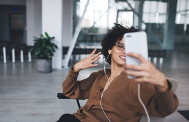 Milenyum Kafkasyalı kadın akıllı telefondan selfie çekerken el kol hareketi yapıyor ve kulaklıkla müzik dinliyor. Modern başarılı kadın yaşam tarzı kavramı. Koltuğunda oturan gözlüklü genç kız.